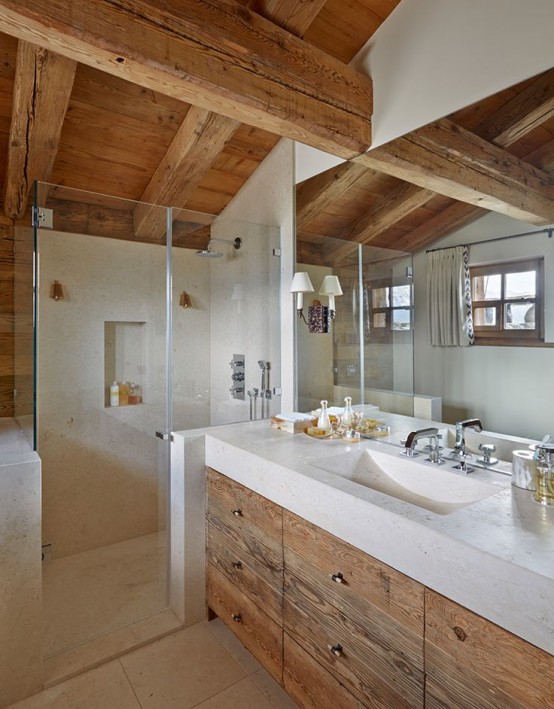 a light-filled chalet bathroom with windows, with a light-stained wood ceiling with beams and a matching vanity, neutral stone on the floor, in the shower