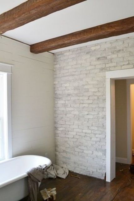 a neutral farmhouse bathroom with whitewashed brick walls, wooden beams, a vintage tub is filled with light and looks cool