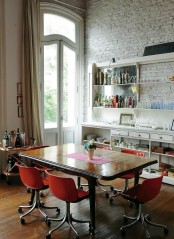 an eclectic dining space with a whitewashed bricl wall, a large buffet and shelves, a refined wooden table and modern red chairs