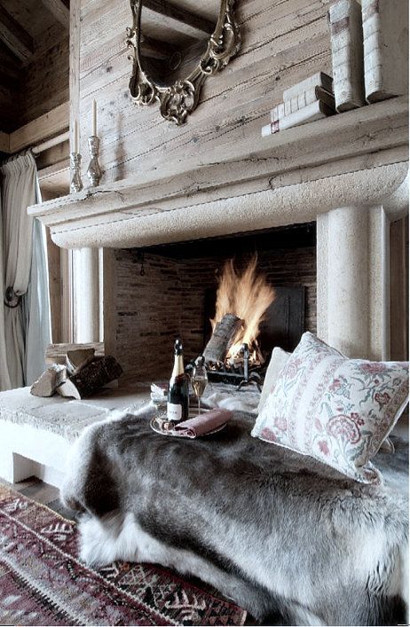 a cool space with an open fireplace clad with stone, with firewood, some animal skins and pillows and a bottle of champagne