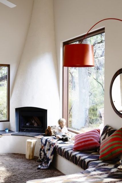 a contemporary space with a real hearth, a windowsill daybed with pillows, a floor lamp in orange and a gorgeous view of the outside