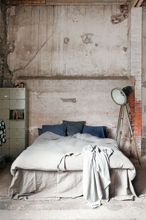 an industrial shabby chic bedroom with rough concrete and brick walls, a neutral bed with neutral bedding, a metal file cabinet for storage and a floor lamp