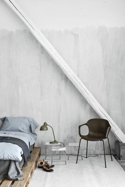 a neutral industrial bedroom with painted walls, a pallet bed with blue bedding, a black chair and a table lamp, small stools