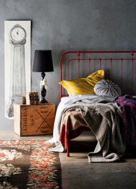 an eclectic bedroom with concrete walls, a red metal bed with bright bedding, a wooden chest as a nightstand and some pretty textiles