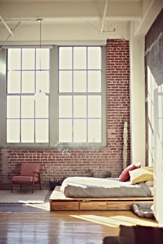 an industrial bedroom with brick walls, double-height windows, a wooden bed with storage drawers and neutral bedding, pipes and metal on the ceiling
