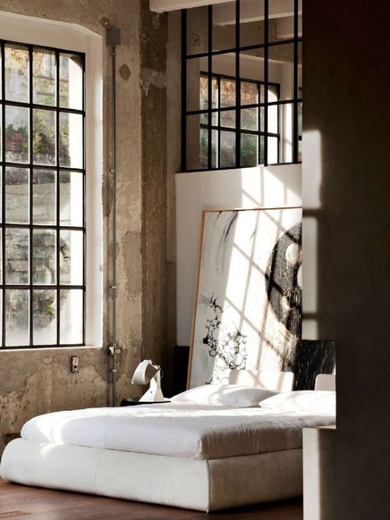 an industrial meets contemporary bedroom with double-height windows, concrete walls and a ceiling, an upholstered bed with neutral bedding, statement artwork