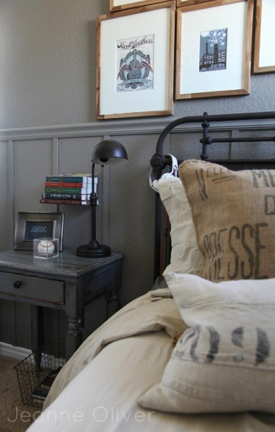 a vintage industrial bedroom with grey walls, a metal bed with printed bedding, a grey nightstand, a black table lamp and a gallery wall