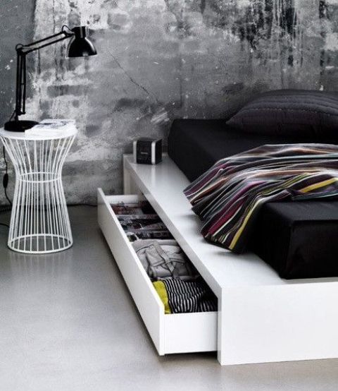 a minimalist meets industrial bedroom with rough concrete walls and floor, a white sleek bed with storage drawers, a white metal nightstand and a black table lamp