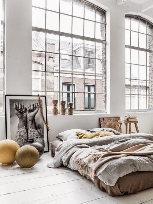 an industrial loft bedroom with oversized arched windows, much natural light and a bed right on the floor, some art is a beautiful space