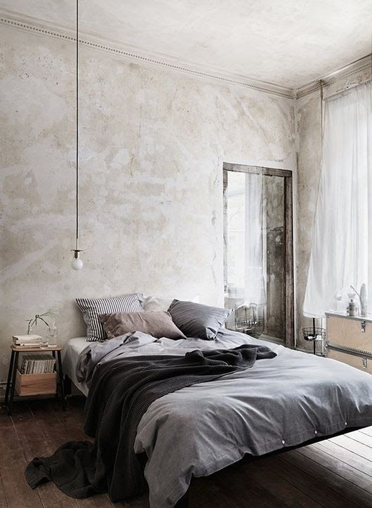 a neutral bedroom with rough walls, a floating bed with grey bedding, a pendant lamp, storage units and some plants is a cool space