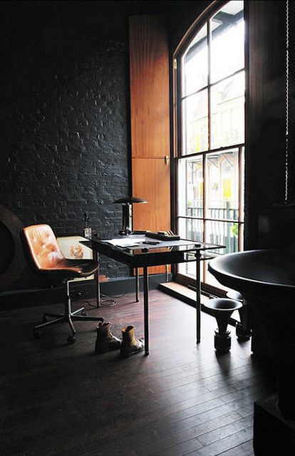 a moody industrial home office with black brick walls, a chic metal and glass desk, a leather chair, a large arched window