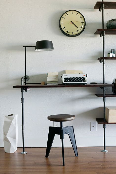 a small industrial working space of a wood and metal shelving unit that includes a desk and a matching stool plus a black table lamp