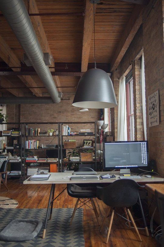 a stylish industrial home office with red brick walls, a cofy desk and a black chair, a metal shelving unit, exposed pipes and metal pendant lamps