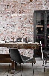 an industrial home office with a red brick wall, a black glass bookcase, a vintage shabby wooden desk and black chairs