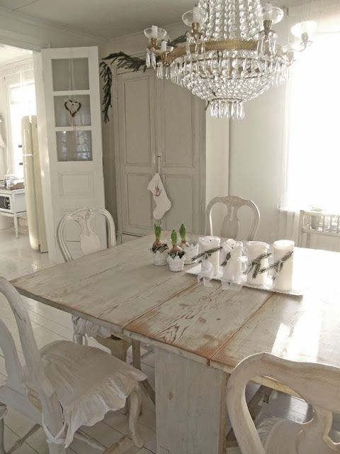 an all-white shabby chic dining room with a shabby storage unit, a shabby chic dining table, white vintage chairs with ruffles and a crystal chandelier