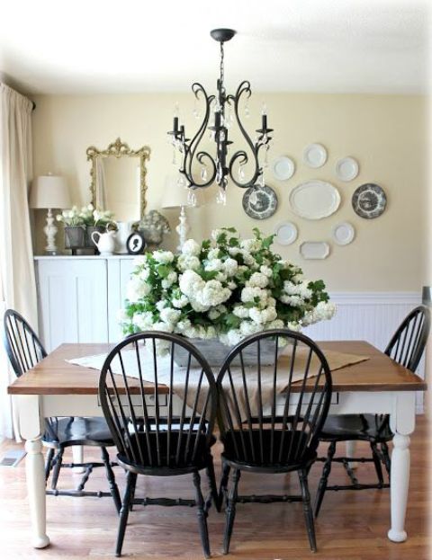 a vintage rustic dining room with light yellow walls and white paneling, a white cabinet, a white dining table with a stained tabletop, matching black chairs, a black vintage chandelier and white blooms and plates on the wall