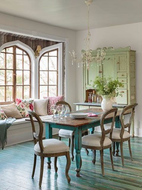 a welcoming vintage dining space with a windowseat with pillows, a shabby dining table with blue legs, a green buffet, matching vintage chairs and greenery