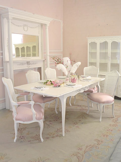 a cute light pink and white vintage dining room with a vintage buffet, a faux fireplace with a mantel, a white vintage table and pink and white matching chairs