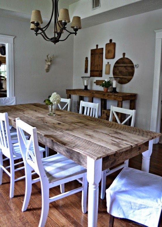 a rustic meets vintage dining zone with a table with a stained tabletop, white chairs, a vintage chandelier, a console table and cutting boards on the wall