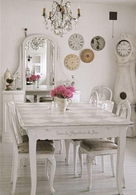 a refined neutral vintage dining zone with a shabby dinign table and matching chairs, a buffet with a mirror, a crystal chandelier and a gallery wall of vintage clocks