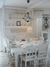 a beautiful all-white vintage dining space with a white table, mismatching white chairs, shelves and a chic crystal chandelier over the table