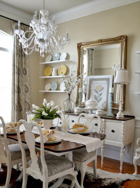 a neutral vintage dining room with tan walls, wall-mounted shelves, a mirror and a buffet, a table with a stained tabletop, white vintage chairs and a white chandelier