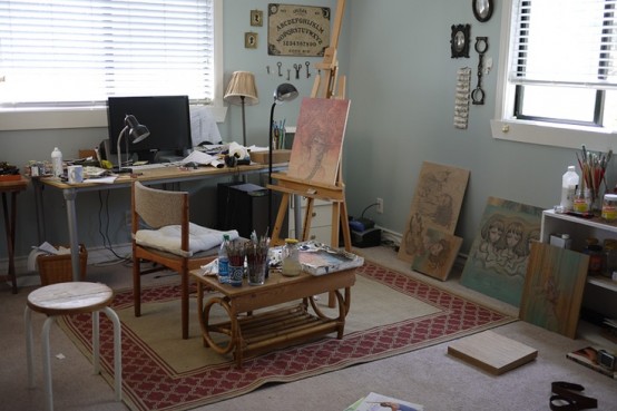 a small and cozy home artist studio with a desk, a chair, an easel, a small table for paints and brushes, some artwork around