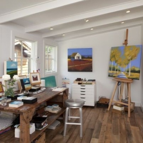 a small and lovely home artist studio with a wooden desk, a stool, a file cabinet, an easel and some bright artwork