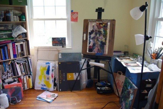 a small and cozy home artist studio with yellow walls, a blue desk, a large shelving unit with books and paints, an easel and some artwork