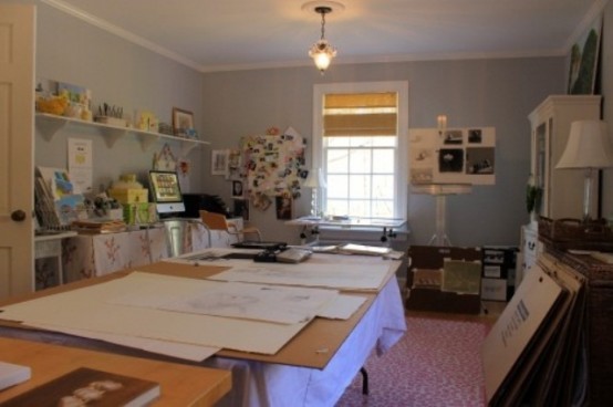 a large home artist studio with grey walls, a large desk, ledges and shelves with artwork, crates, boxes and paints and paper