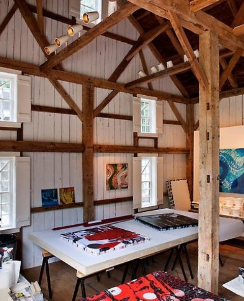 a modern farmhouse artist home studio with wooden beams and pillars, ledges, shelves and a large table, chairs and benches