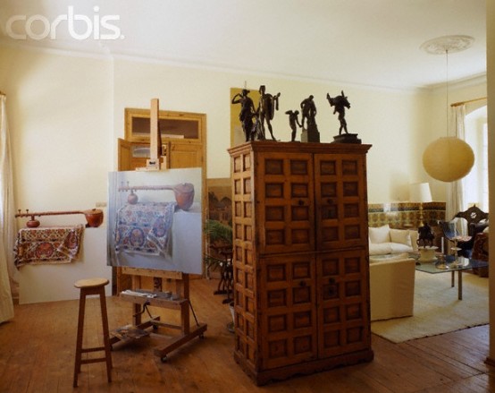 a welcoming and refined home artist nook with an easel, a vintage storage cbainet, some statuettes on top, a tall stool is a cool space