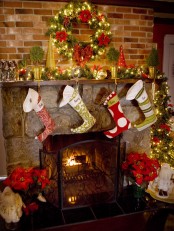 a traditional Christmas mantel with en evergreen garland with lights, potted mini trees and a bold Christmas wreath with red blooms and stockings hanging over the fireplace