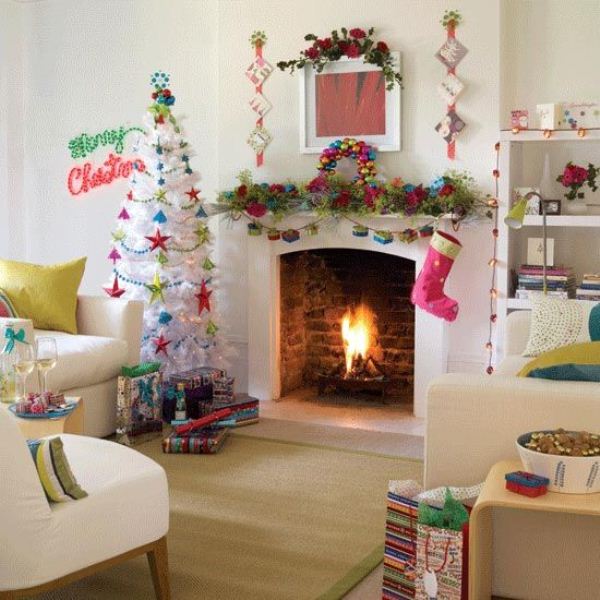a bright Christmas mantel with a greenery garland, bold faux blooms and a matching gift box garland, a pink stocking and a white Christmas tree with colorful ornaments