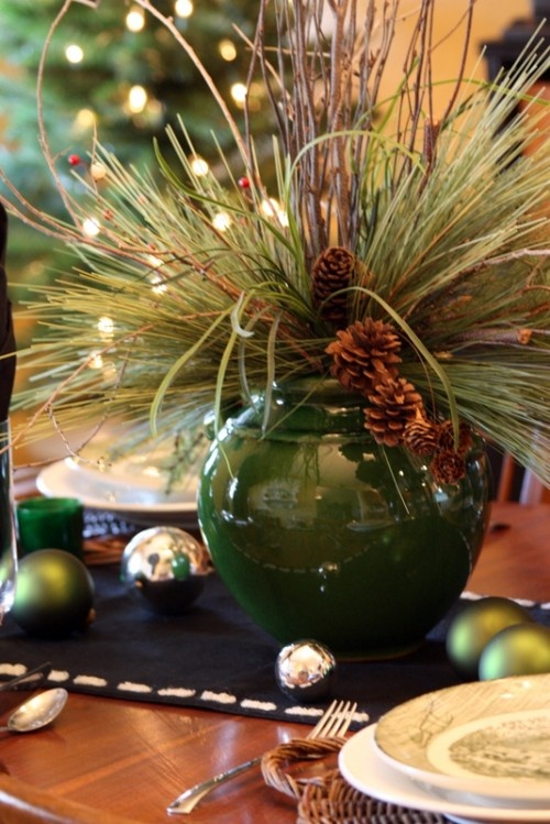a rustic Christmas tablescape with a black runner, green and silver ornaments, an evergreen and pinecone centerpiece plus woven placemats