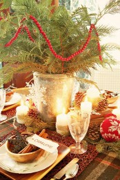a rustic Christmas tablescape with a plaid tablecloth, a red woven placemat, pinecones, candles, evergreens and a wooden charger