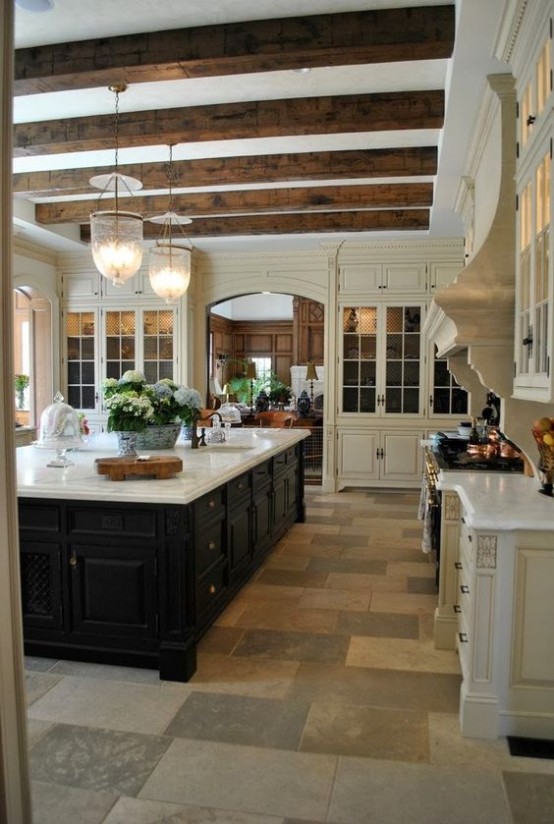 a refined vintahe kitchen in warm neutral tones, with a dark kitchen island, dark wooden beams and pendant lamps is all chic