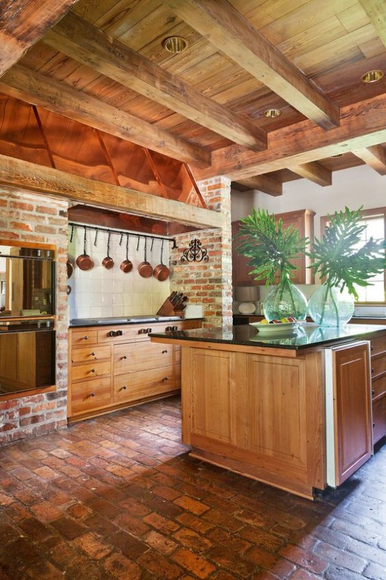 a cozy warm-tone kitchen with wooden cabinetry, brock walls, black stone countertops, wooden beams on the ceiling to add interest and an architectural element to the space