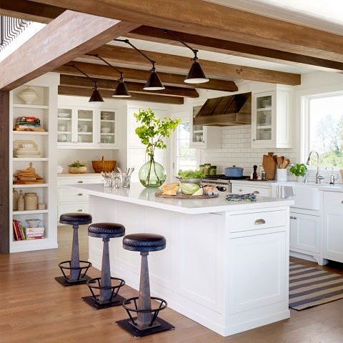 a neutral modern farmhouse kitchen with white cabinetry, wooden beams on the ceiling with black sconces, black stools and greenery for a fresh touch
