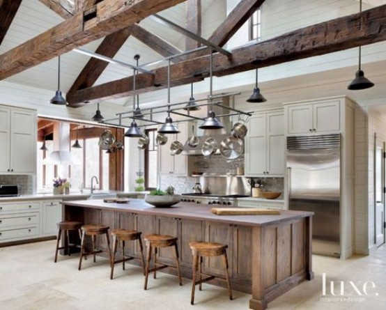 a neutral modern farmhouse kitchen with neutral cabinetry, a dark heavy kitchen island, with dark wooden beams, pendant lamps attached to them is a welcoming space