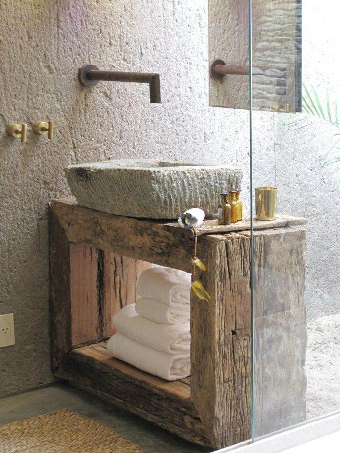 a wabi sabi bathroom with a wooden console, white stone walls and a matching sink plus metal fixtures