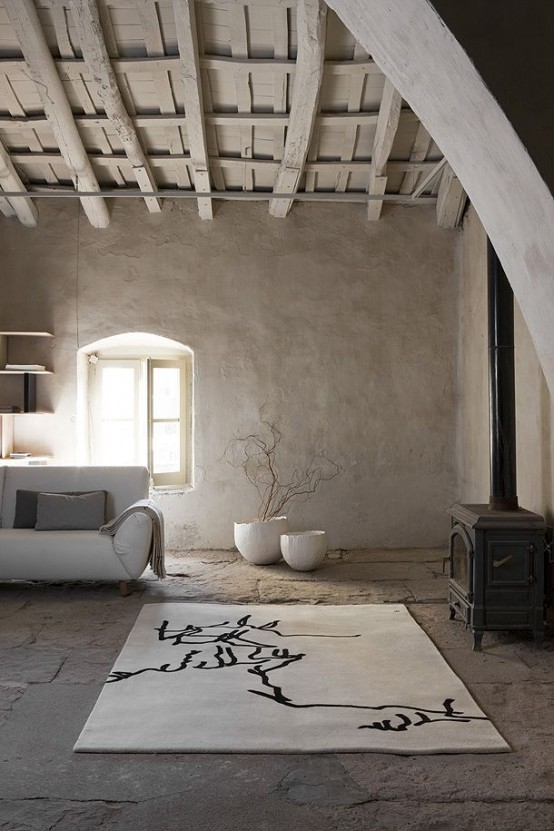 a neutral living room with a wooden roof and beams, a rough stone floor, rough concrete walls and minimal furniture