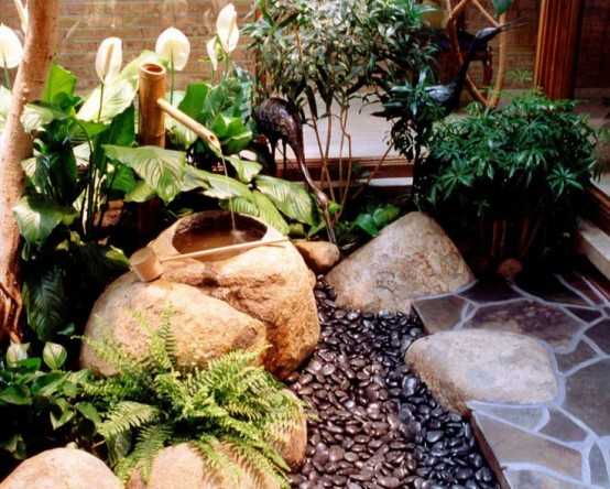 a mini Japanese garden with dark pebbles, a traditional bamboo fountain, large rocks, greenery and ferns