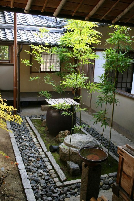 un patio japonés con guijarros, rocas, musgo, una piedra grande, un cuenco con una cubierta de bambú y algo de vegetación