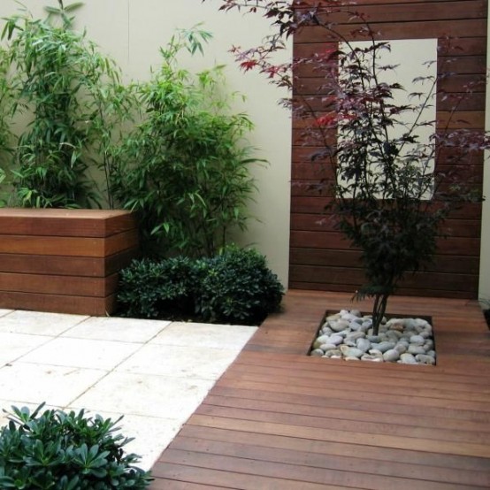 una plataforma de madera contemporánea con un solo árbol con guijarros, vegetación y una pared de madera