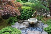 a small Japanese garden with pebbles, rocks, a bamboo fountain, greenery and trees around