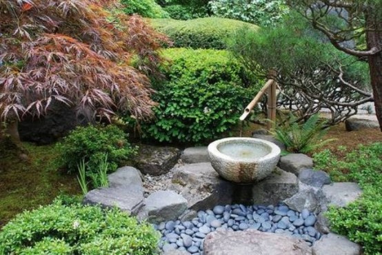 un pequeño jardín japonés con guijarros, rocas, una fuente de bambú, vegetación y árboles alrededor