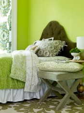 a bold green bedroom with a dark green upholstered bed, green and white bedding, a grey upholstered stool and a printed pillow and some printed curtains