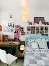 a small and pretty girl’s room with a bed with printed bedding, box-shaped shelves, a bright gallery wall, a desk and a creamy chair, paper lights