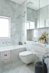 a neutral contemporary bathroom clad with marble tiles, with a tub, a toilet and a large mirror cabinet on the wall
